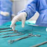 Nurse hand taking surgical instrument for group of surgeons at background operating patient in surgical theatre. Steel medical instruments ready to be used. Surgery and emergency concept